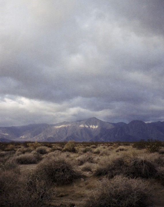 A desert landscape with scrub brush in the foreground and mountains under a cloudy sky.