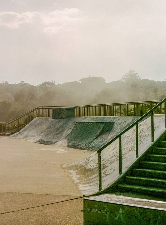A skatepark with ramps and stairs is shrouded in morning fog, with trees and houses visible in the background.