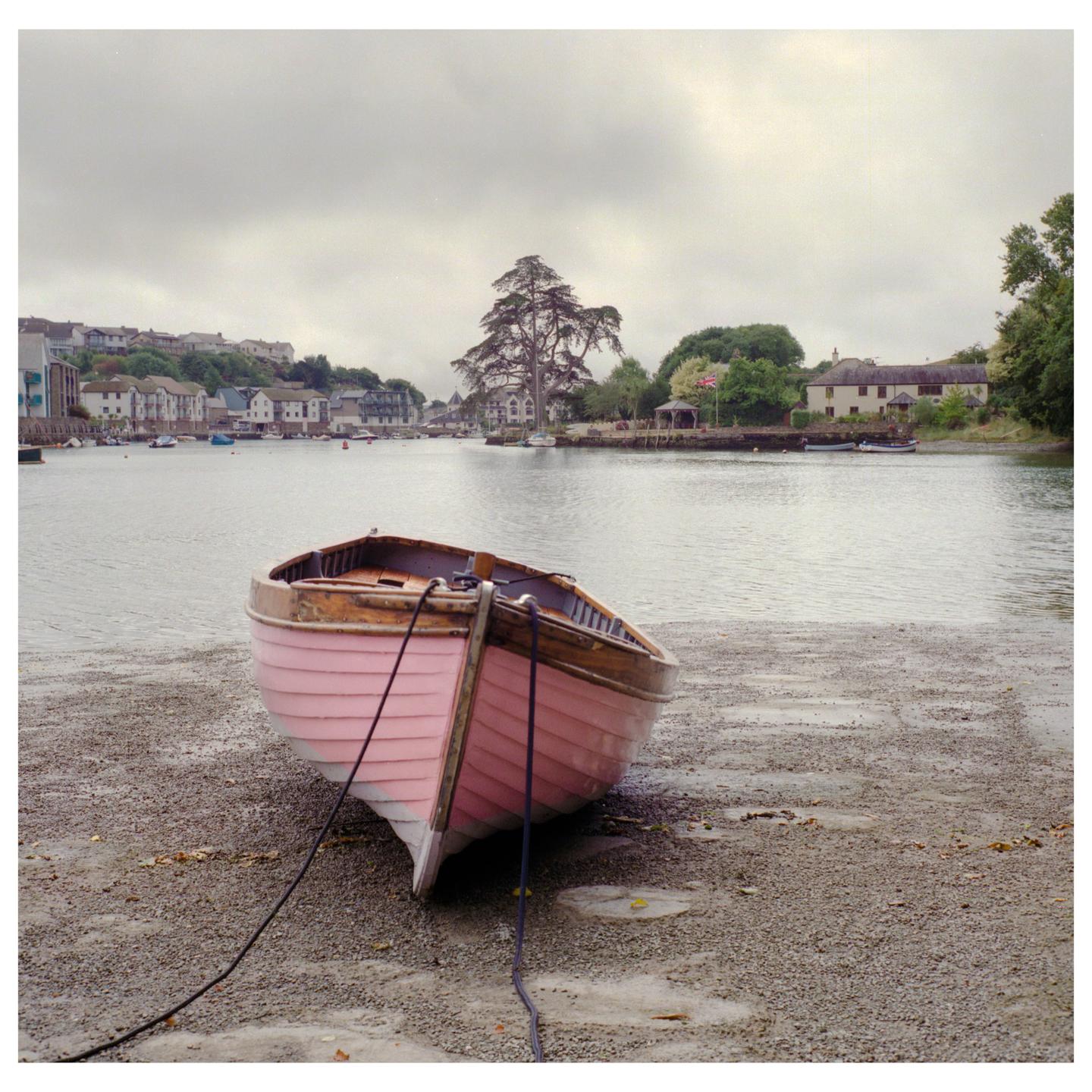 A pink rowboat rests on a rocky shore with a town and river in the background under a cloudy sky.