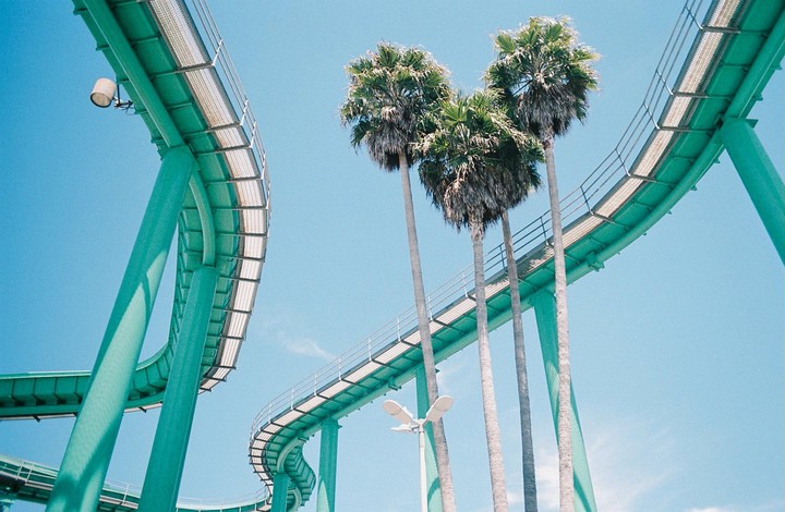 A teal roller coaster track curves through the sky with palm trees in the background.