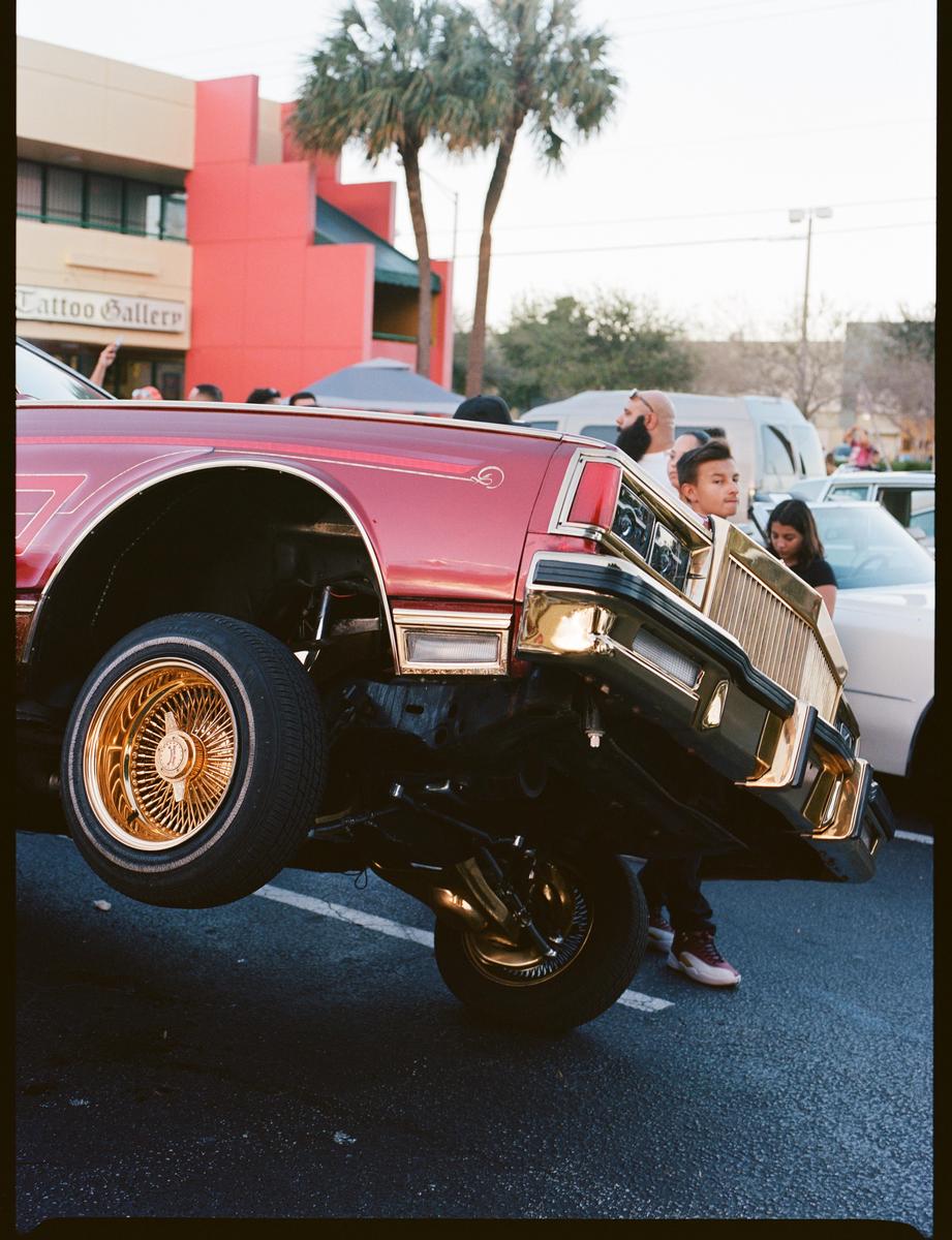 A red lowrider with gold accents is bouncing on its hydraulic suspension, lifting its front wheel off the ground, while people watch in the background.