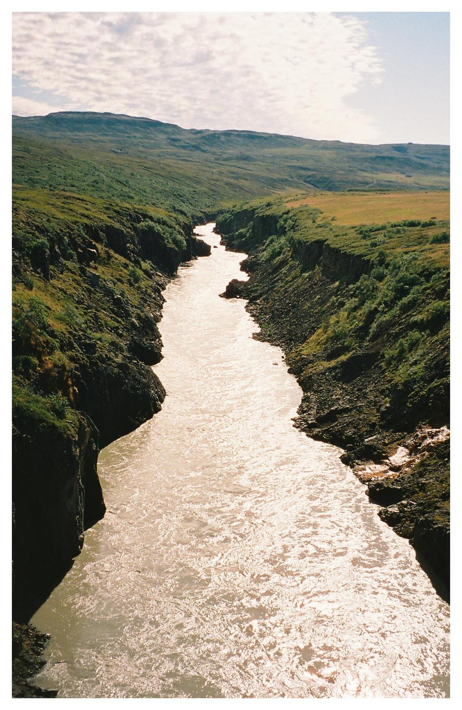 A river flows through a canyon with green hills on either side under a cloudy sky.