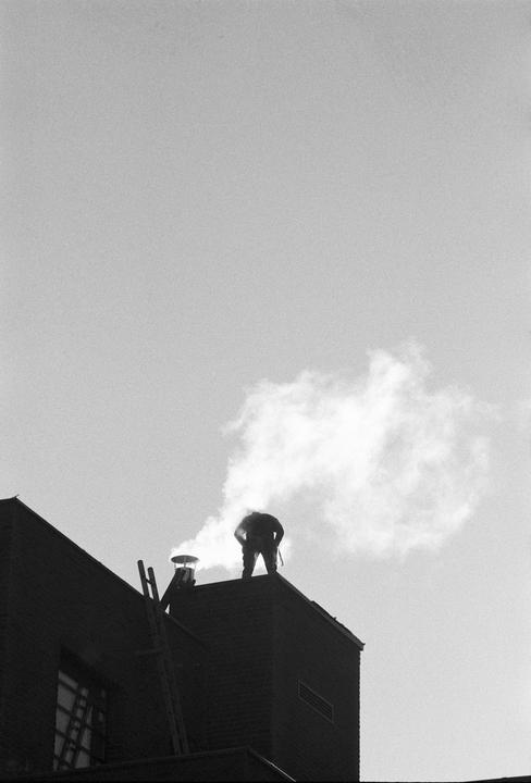 A man stands on a rooftop next to a smoking chimney, with a ladder leaning against the building.