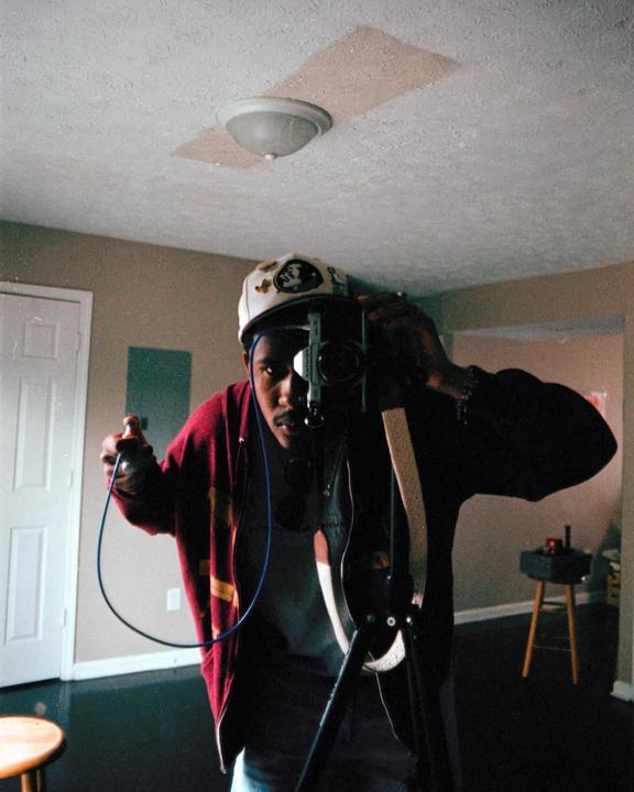 A man wearing a cap and a red jacket takes a self-portrait with a camera on a tripod.