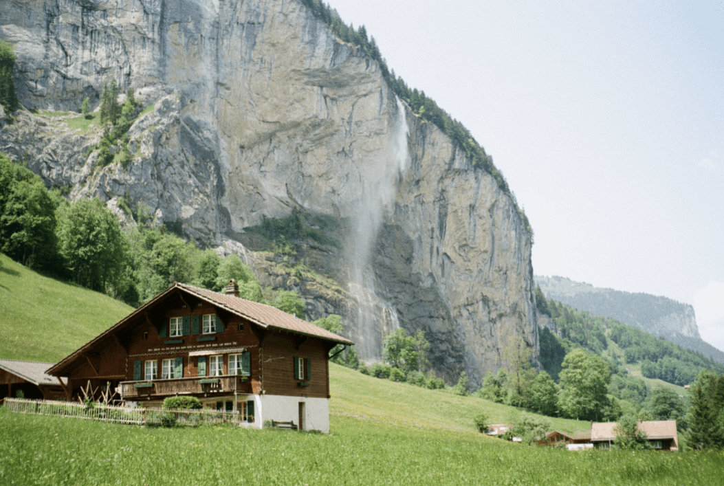 A wooden house with green shutters sits in a grassy field with a large waterfall cascading down a cliff face in the background.