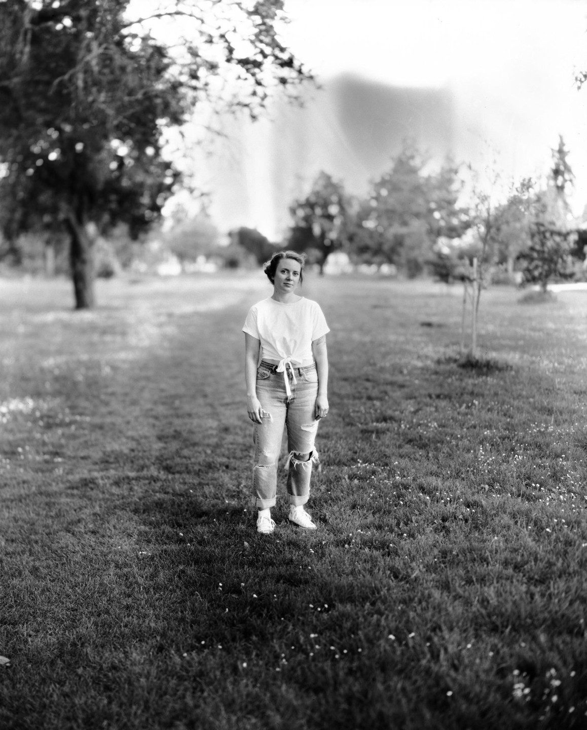 A woman stands in a grassy field with trees in the background.