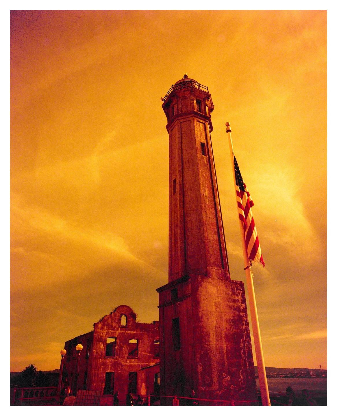 A tall tower stands next to a flagpole with an American flag, with a ruined building in the background under an orange sky.