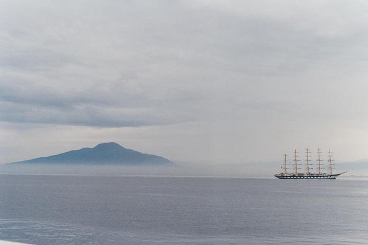 A large sailing ship with multiple masts floats on the water with a hazy mountain in the background under a cloudy sky.