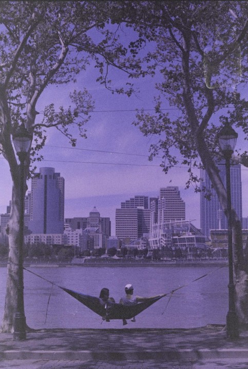Two people relax in a hammock by a river with a city skyline in the background.