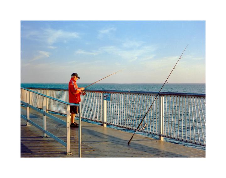 A man in a red jacket and baseball cap is fishing from a pier over the ocean.