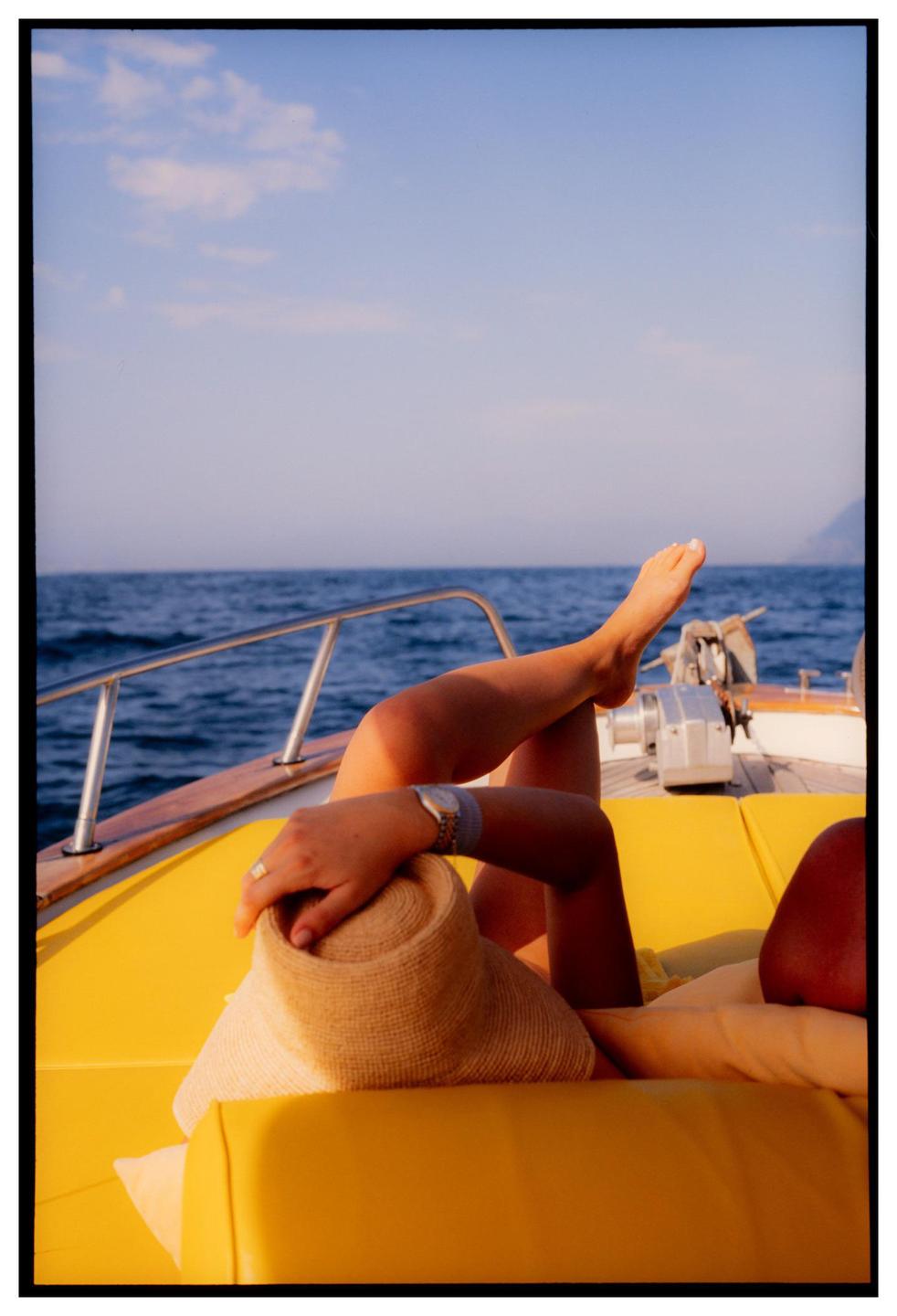 A woman relaxes on a boat with her legs crossed and a straw hat on her lap.