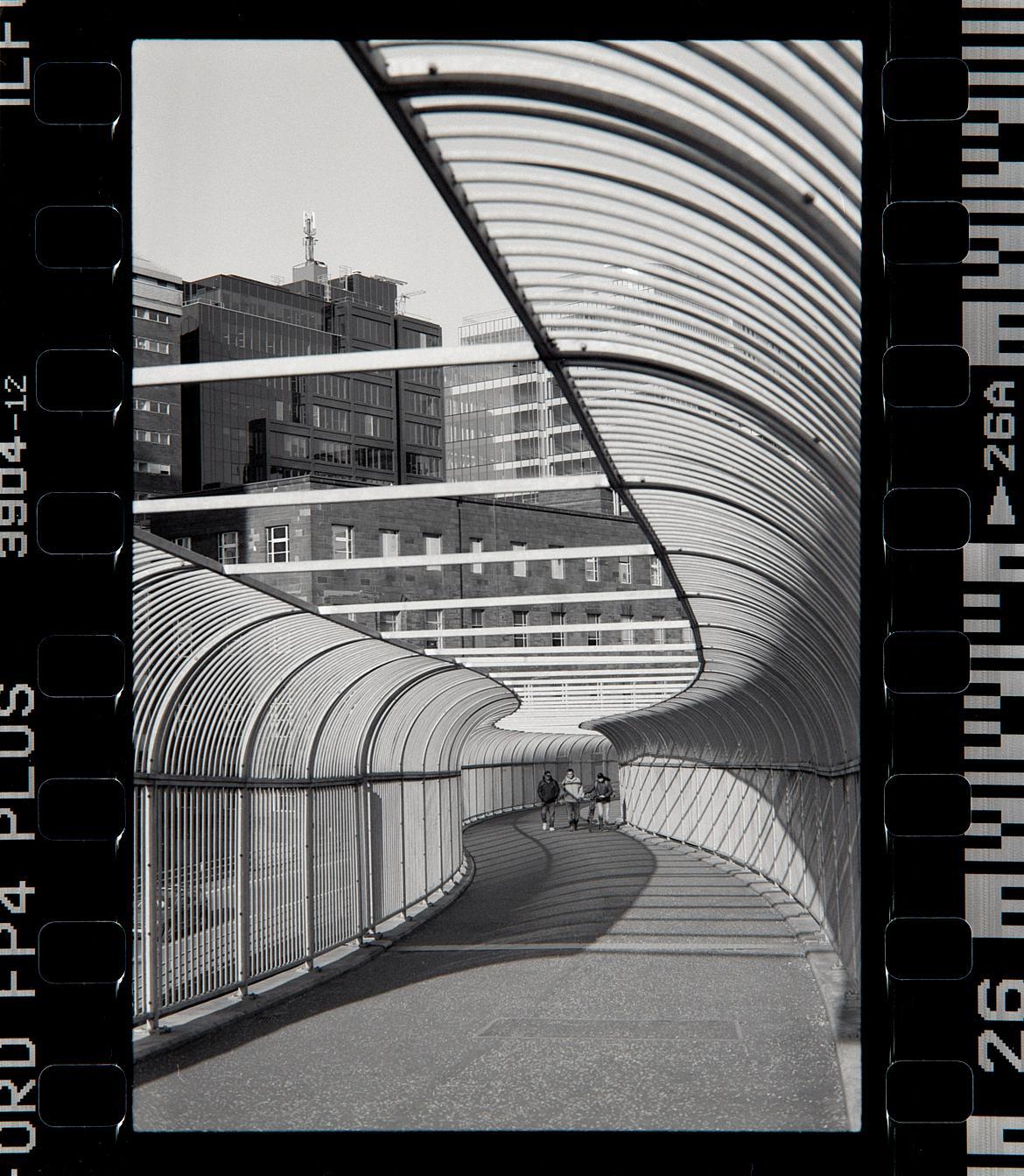 Three people walk through a curved, covered walkway with buildings in the background.
