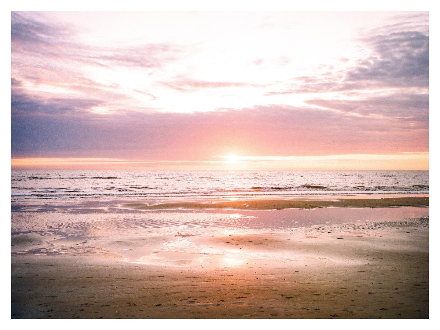 The sun sets over a beach with soft waves and colorful clouds.