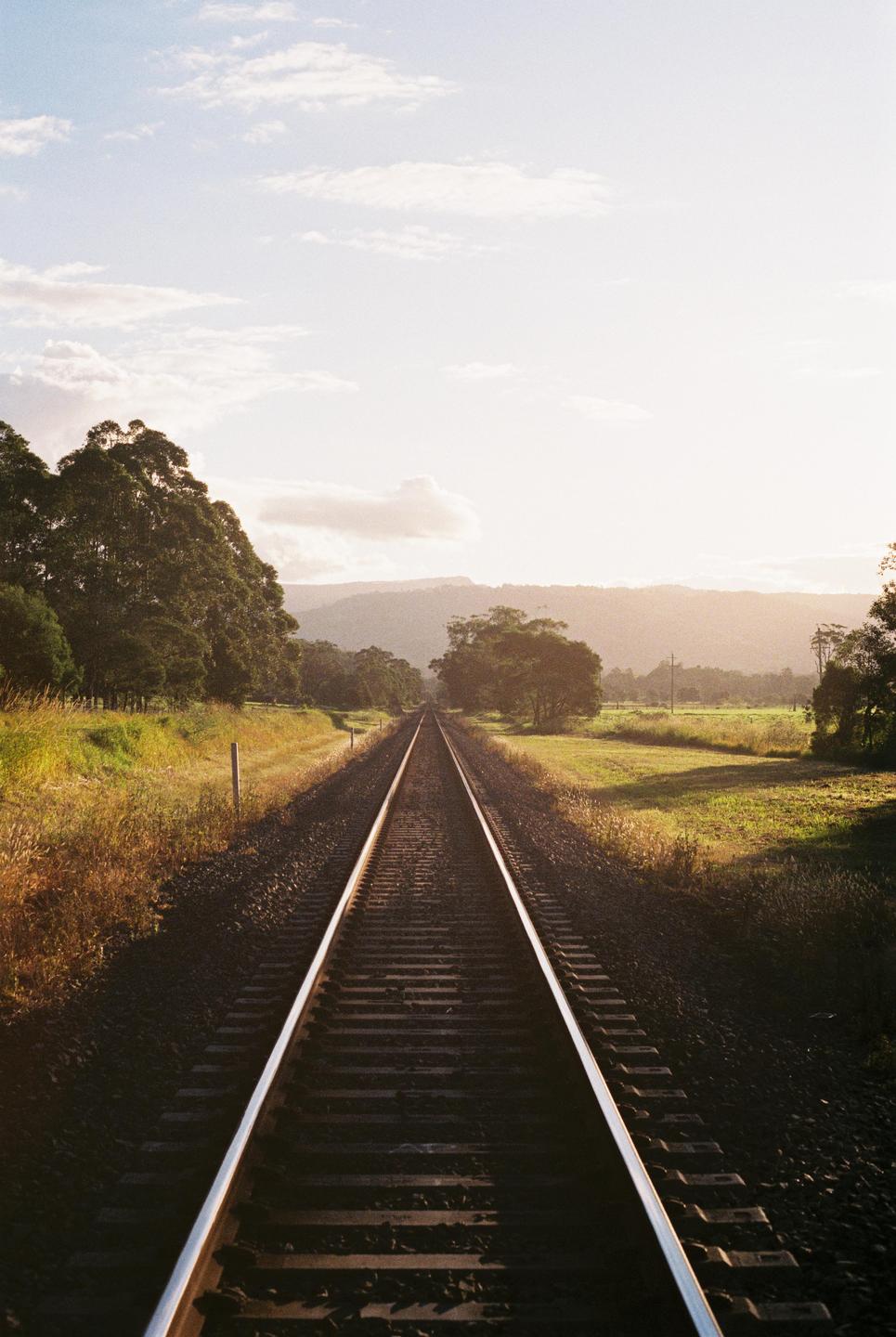 A long railway track stretches into the distance, flanked by trees and fields under a bright sky.