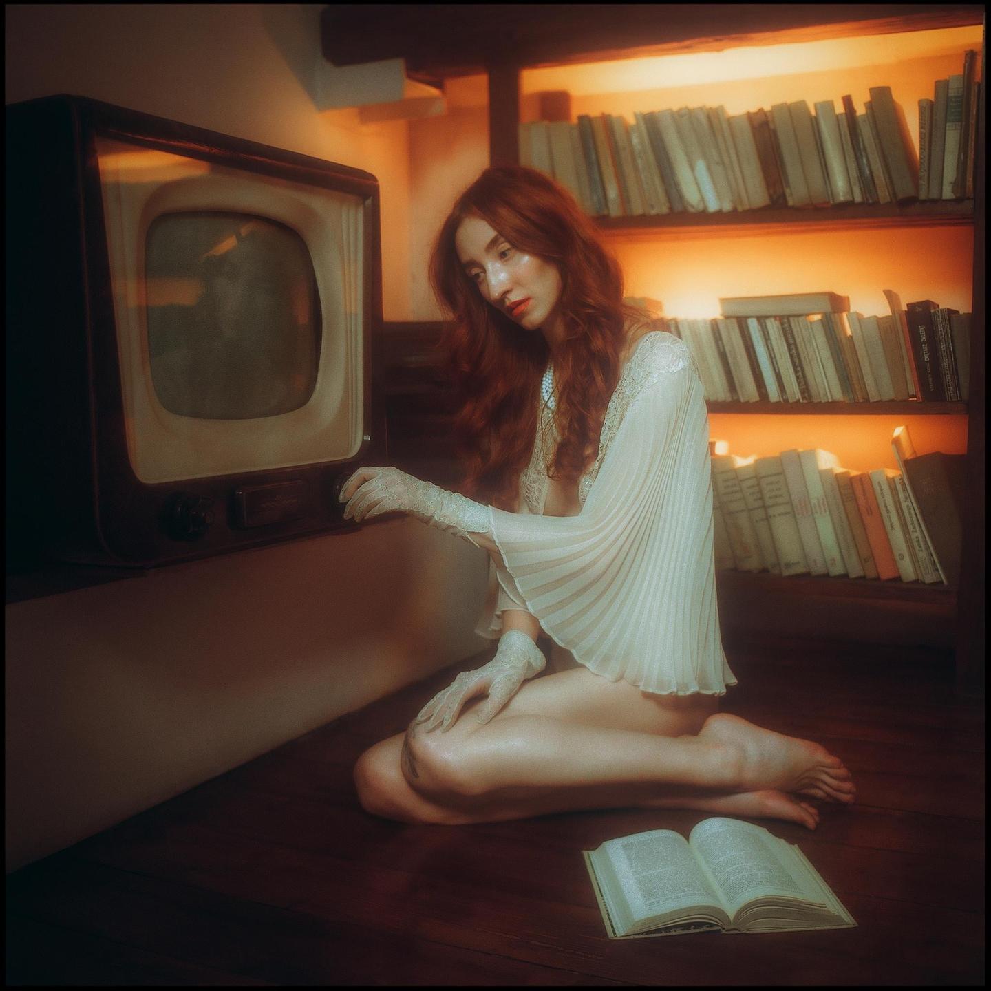A woman with long red hair kneels on a wooden floor next to an old television and an open book, with a bookshelf in the background.