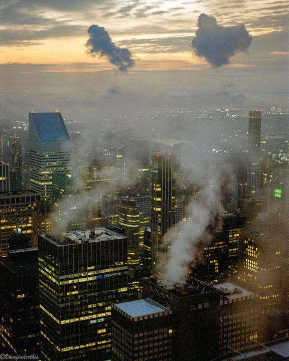 An aerial view of a city at dusk shows illuminated buildings and steam rising from rooftops, with a hazy sky above.