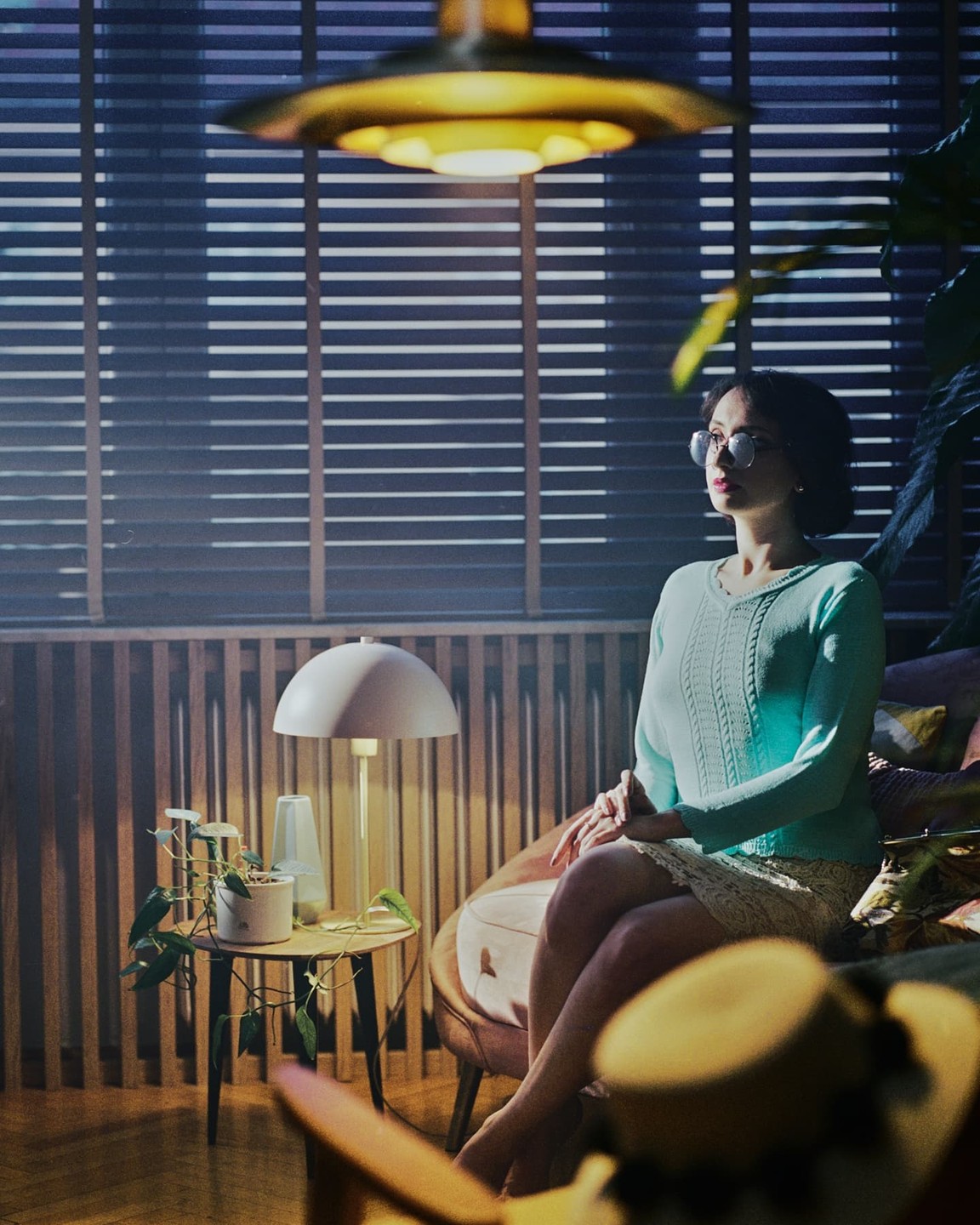 A woman in glasses sits in a chair next to a small table with a plant and a lamp, looking towards a window with blinds.