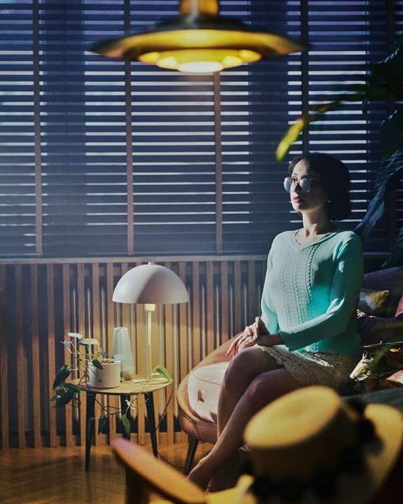 A woman in glasses sits in a chair next to a small table with a plant and a lamp, looking towards a window with blinds.