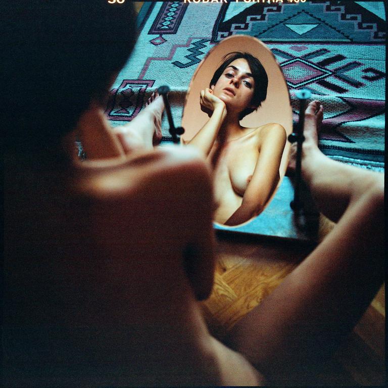 A woman is reflected in a mirror, with her back to the viewer, sitting on a wooden floor with a patterned rug.
