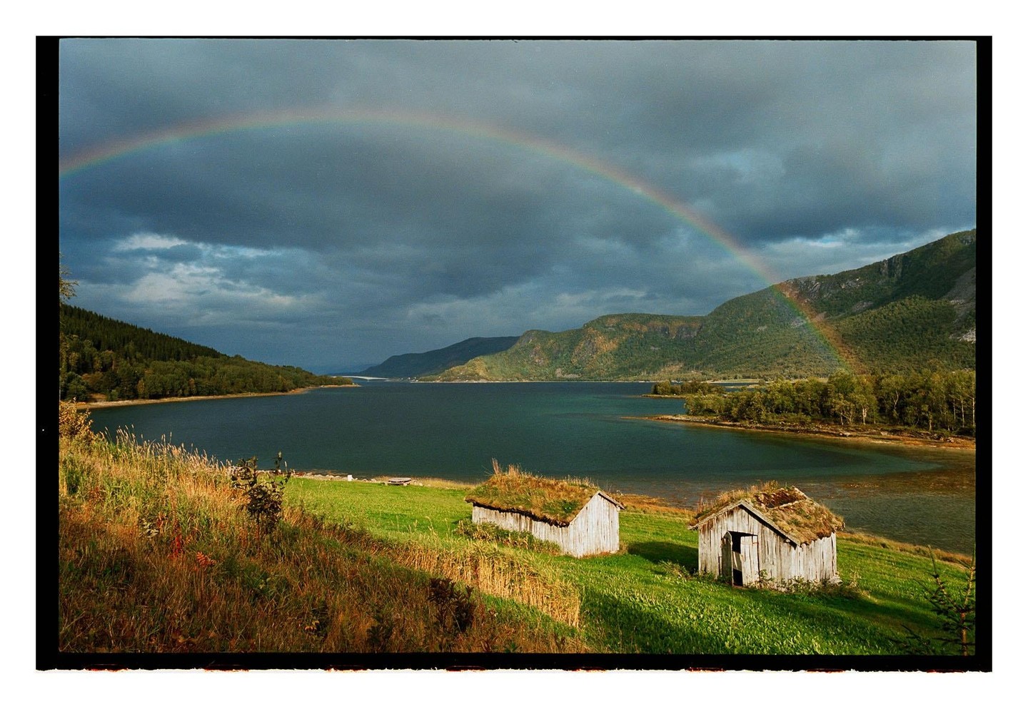 Two small wooden sheds with grass roofs sit on a green hillside overlooking a lake with a rainbow arcing over it.