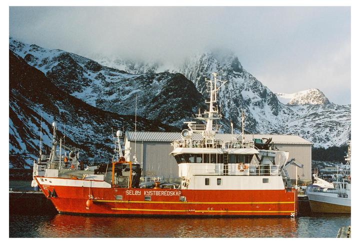 A red and white fishing boat is docked in a harbor with snow-covered mountains in the background.