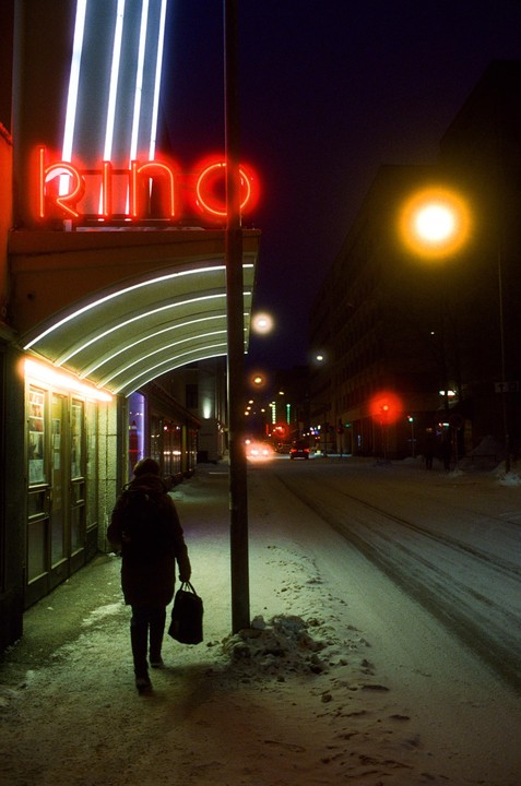 A person walks on a snowy sidewalk past a building with a neon "Kino" sign at night.