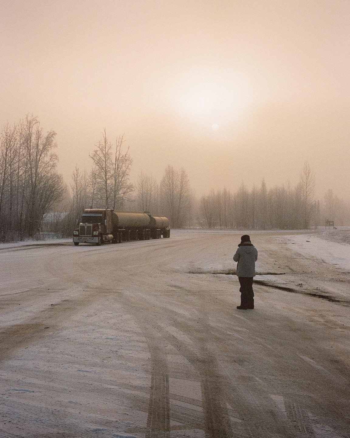 A person stands on a snowy road with a tanker truck and bare trees in the background under a hazy sky.