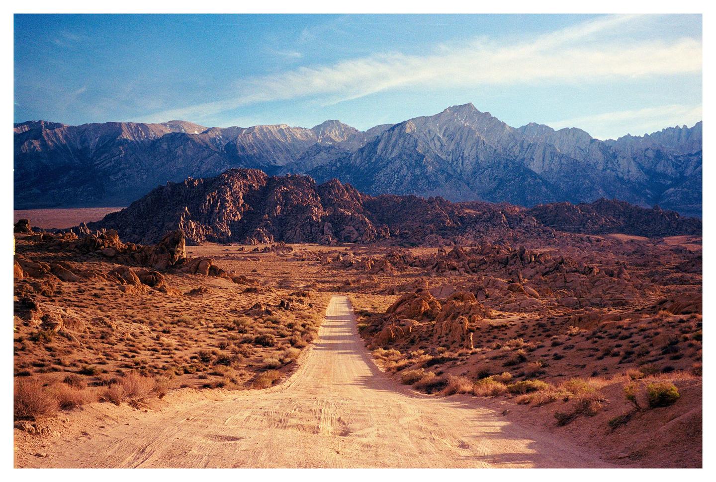 A dirt road leads through a desert landscape towards mountains under a blue sky.