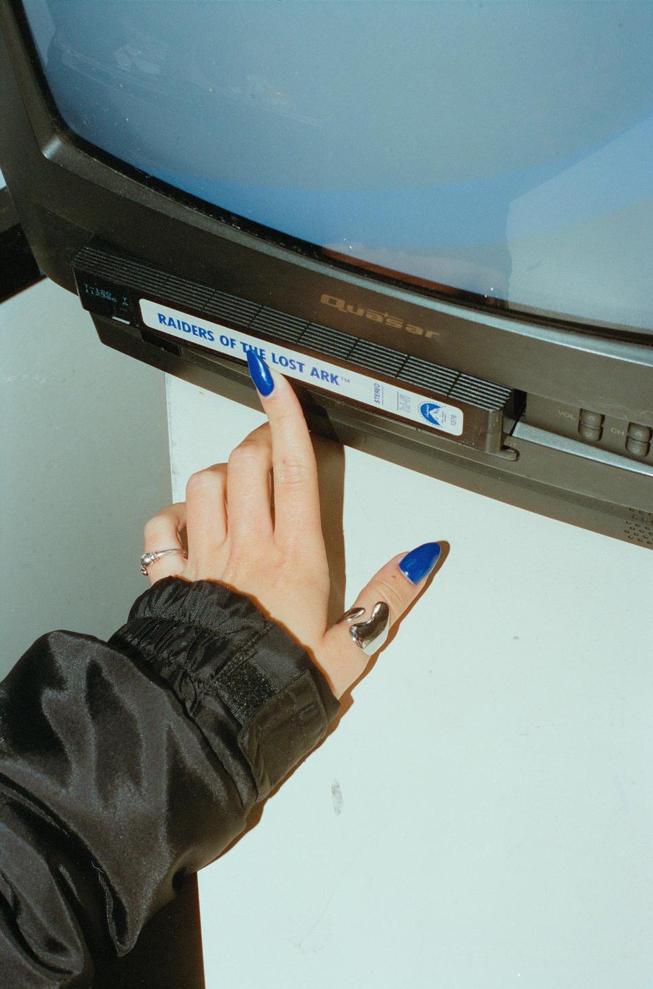 A hand with blue nails inserts a VHS tape labeled "Raiders of the Lost Ark" into a television.