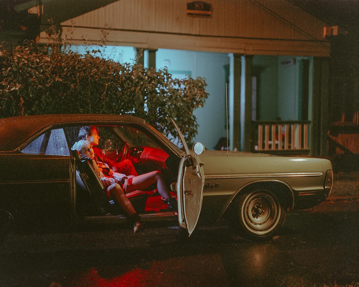 A couple sits in a vintage car at night with the door open, illuminated by red light inside the vehicle.