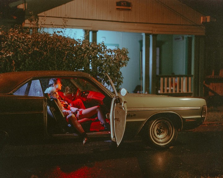 A couple sits in a vintage car at night with the door open, illuminated by red light inside the vehicle.
