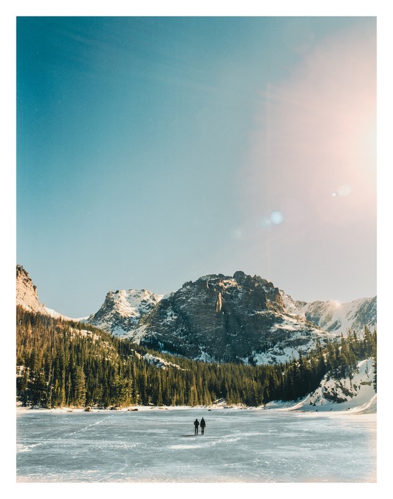 Two people walk across a frozen lake surrounded by snow-capped mountains and a forest.