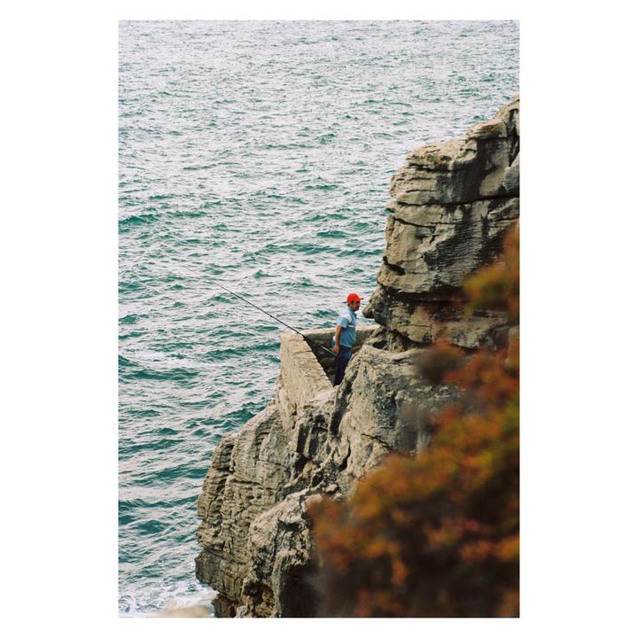 A man in a red hat fishes from a rocky cliff overlooking the ocean.