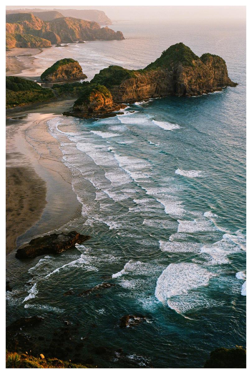 Waves crash on a sandy beach next to a rocky, green coastline under a bright sky.