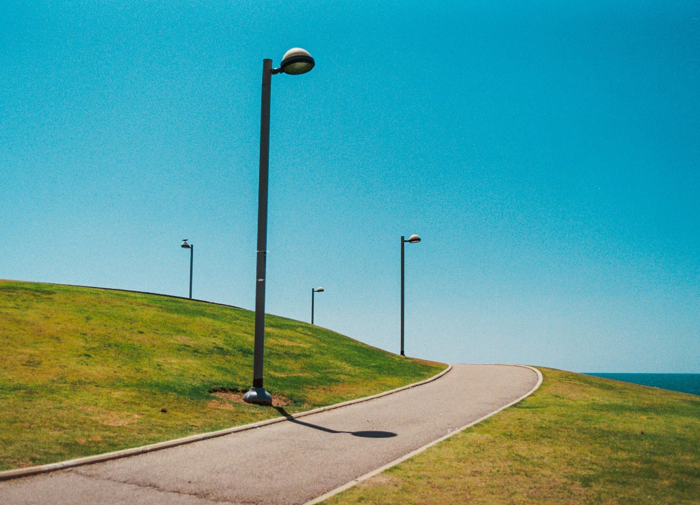 A winding path with streetlights on a grassy hill leads towards the ocean under a clear blue sky.