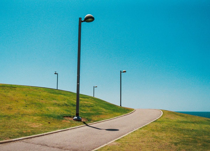 A winding path with streetlights on a grassy hill leads towards the ocean under a clear blue sky.