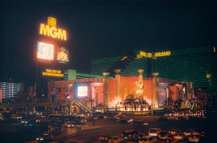 A large building with bright signs and a statue of a lion is visible at night, with many cars on the street in front of it.