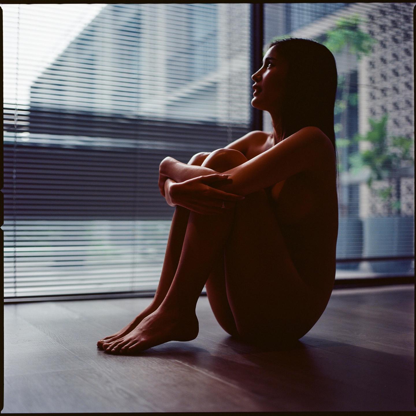 A woman sits on the floor with her knees drawn up, looking out a window with blinds.