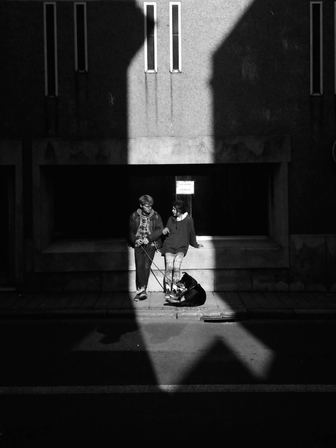 A couple and their dog stand on a sunlit sidewalk next to a building with shadows cast across it.