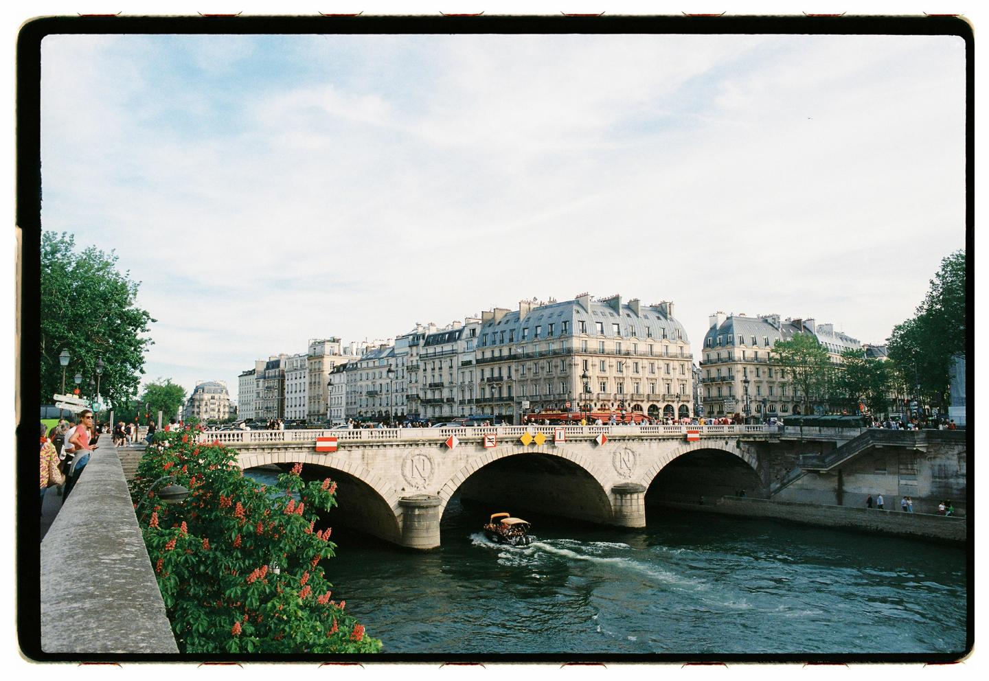 A boat travels on the river Seine with a bridge and buildings visible in the background.