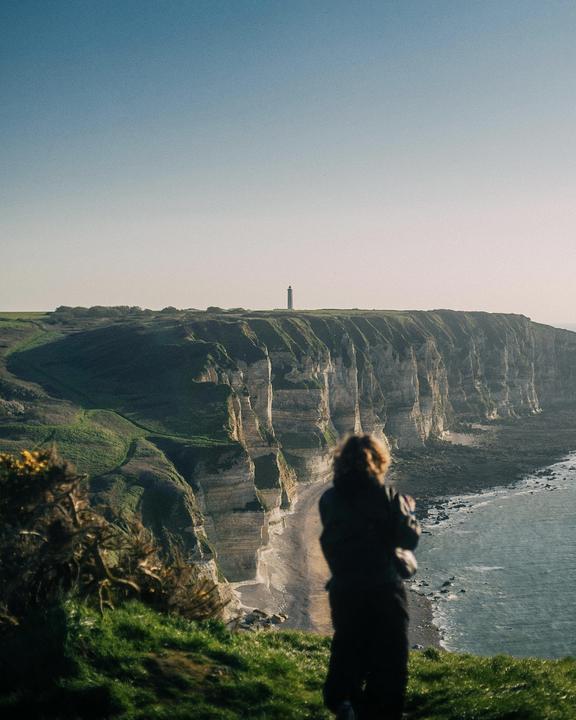 A person stands on a grassy cliff overlooking a beach, ocean, and distant lighthouse.