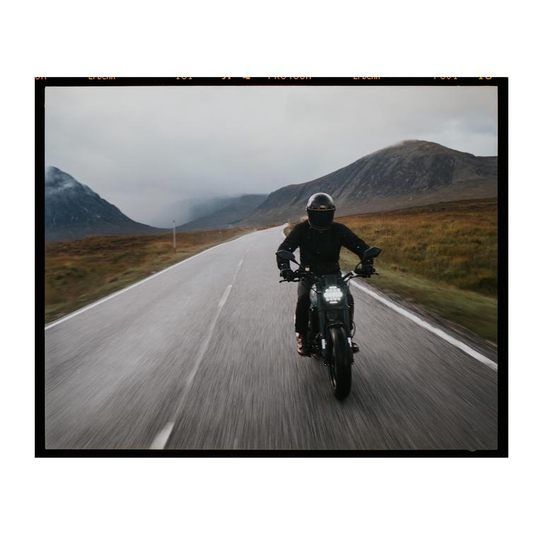 A person on a motorcycle rides down a road with mountains in the background.