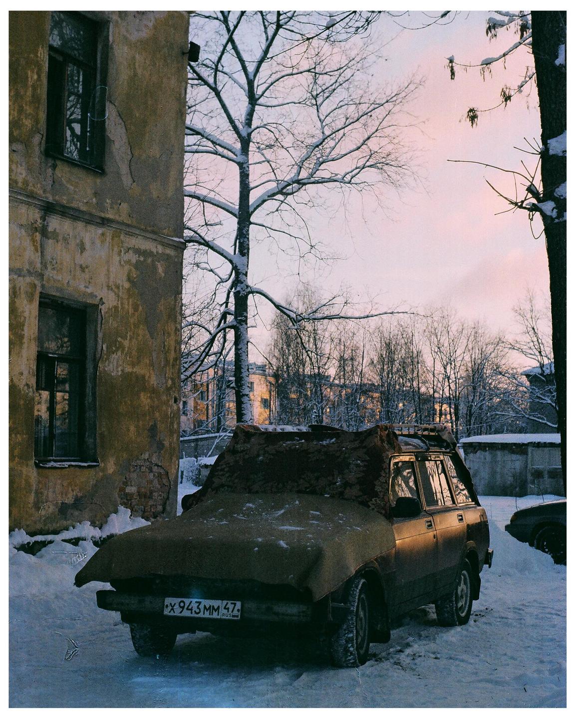 A car covered with a tarp is parked in the snow next to a building with a bare tree in the background.