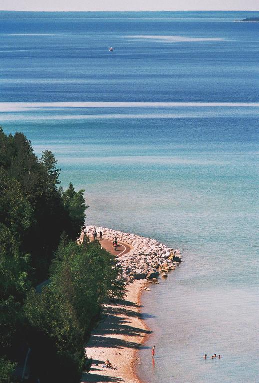 A high-angle view shows a winding path along a rocky and sandy shore with people swimming and walking, and a boat in the distance on the blue water.