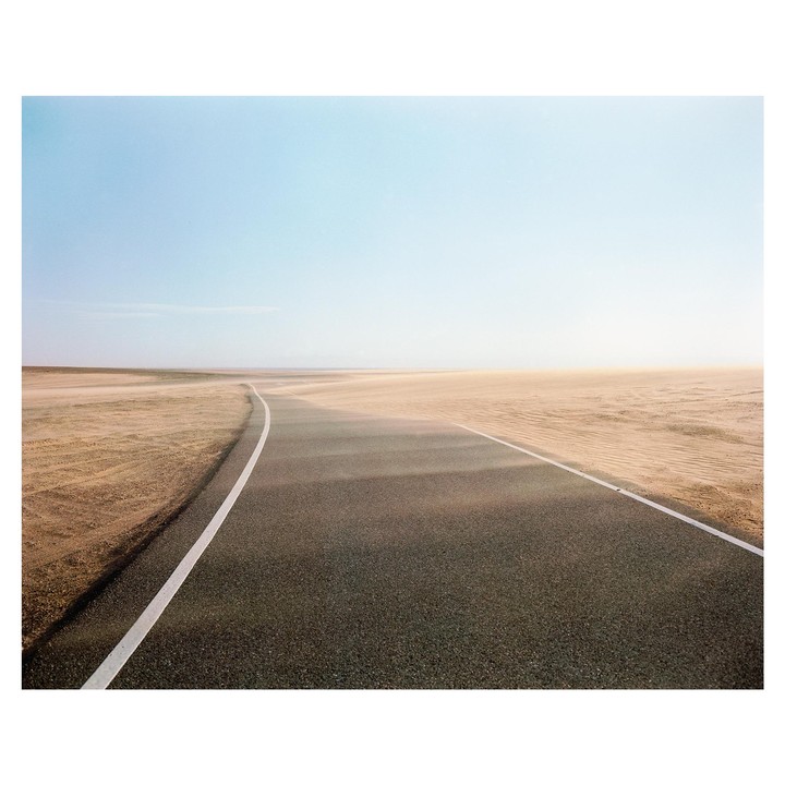 A paved road with white lines curves through a sandy desert under a clear sky.
