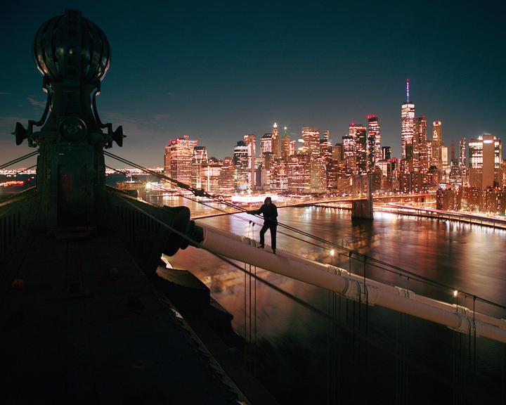 A person stands on a bridge cable overlooking a city skyline at night.