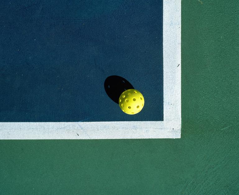 A yellow pickleball with a shadow sits on the corner of a blue and green court with white lines.