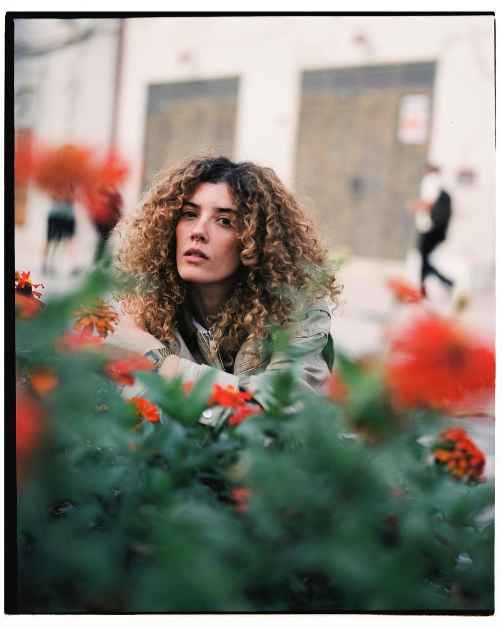 A woman with curly hair looks directly at the viewer from behind a bush with red flowers.