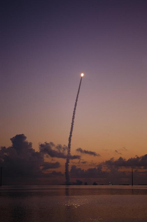 A rocket launches into the sky, leaving a trail of smoke over a body of water at sunset.