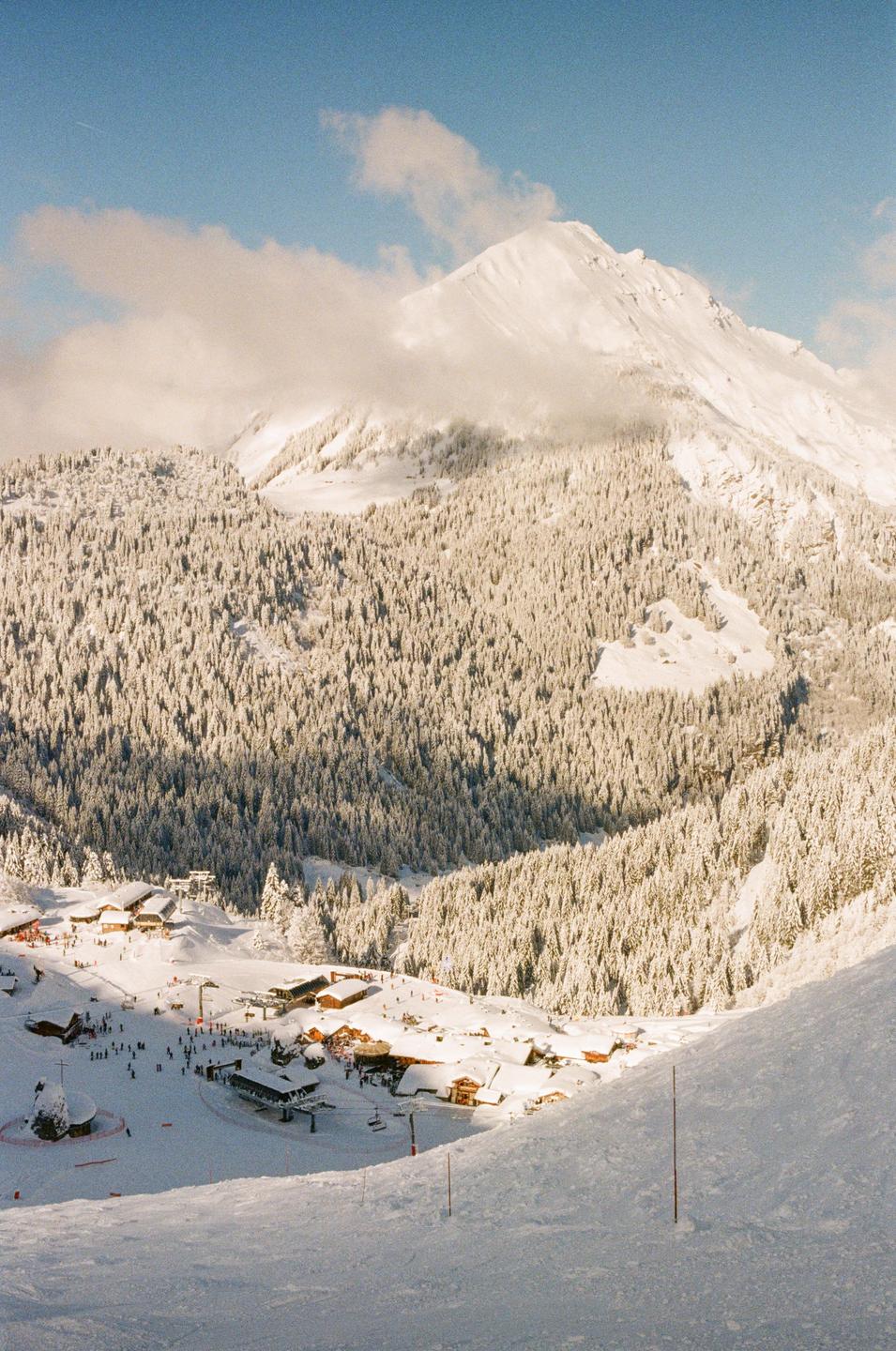 A snow-covered mountain landscape features a ski resort village nestled among pine trees, with a large peak rising in the background.